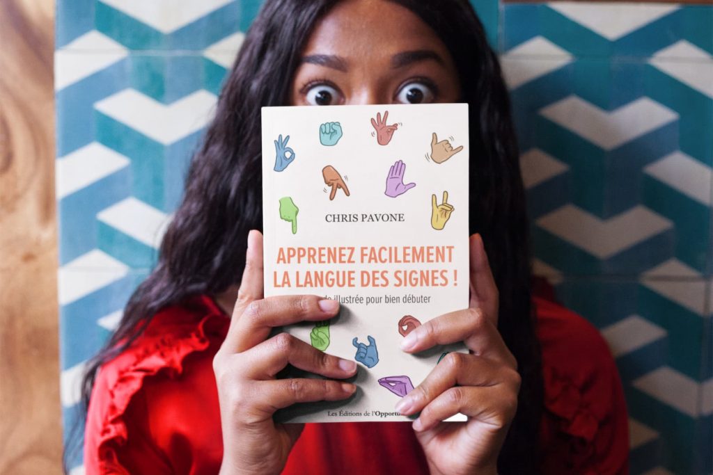Une jeune femme tient dans ses mains, dissimulant sa bouche, le livre de Chris Pavone « Apprenez Facilement la Langue des Signes ! – Méthode illustrée pour bien débuter »